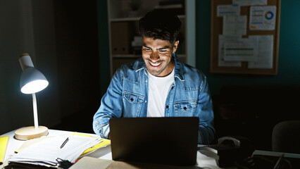 Obraz premium A smiling young adult man works late in an office, illuminated by a lamp in a dark room, with paperwork and a laptop.