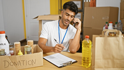 A young man volunteers at a food donation center, organizing supplies and taking notes while on the phone.