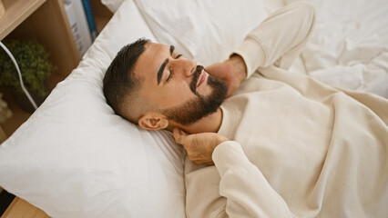 A young man with a beard appears in pain holding his neck while lying in bed indoors, giving a sense of discomfort.