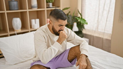 A thoughtful man with a beard sits on a bed in a cozy bedroom, clutching a tissue.