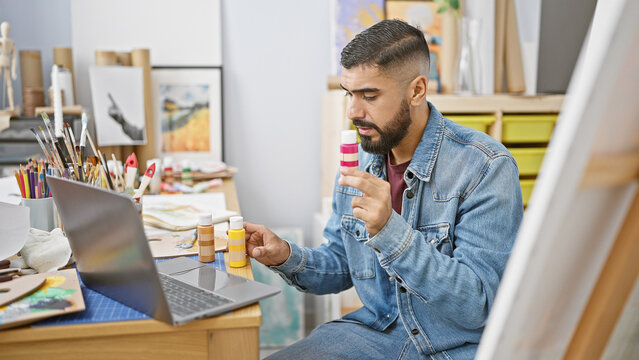 A bearded man examines paint bottles during a creative session in a vibrant art studio, with a laptop nearby.