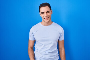 Young hispanic man standing over blue background winking looking at the camera with sexy...