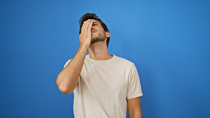 A stressed young man with a beard stands against a plain blue background, visibly upset or frustrated.