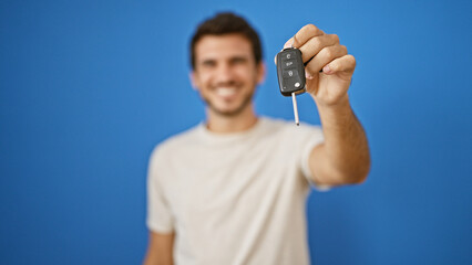 A smiling young hispanic man holding car keys against a blue background, portraying a sense of...