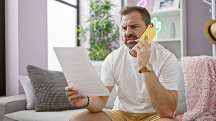 Mature hispanic man with grey hair reading a document and talking on a yellow phone in a modern living room.