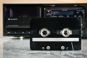 Black audio cassette with empty white space for notes in front of cassette deck on marble shelf with reflection