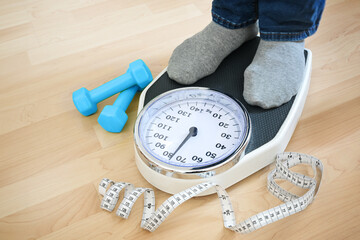 Feet of a man in gray socks standing on a scale to check the weight after fitness training, blue dumbbells and a measuring tape lying nearby on a wooden floor, copy space