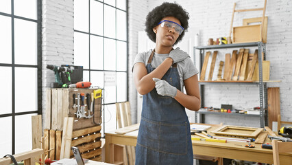 Black woman in safety glasses at a carpentry workshop holds shoulder implying injury indoors.