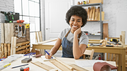 Smiling african american woman taking notes in a sunny carpentry workshop, surrounded by tools and...