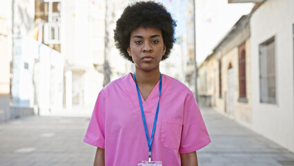 Portrait of a confident young african american female healthcare worker outdoors, wearing a pink scrub and an id badge.