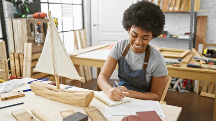 A smiling african american woman carpenter writing in a notebook in a woodwork studio workshop.