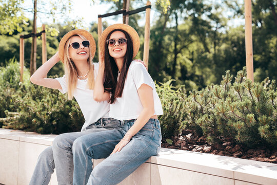 Two young beautiful smiling hipster female in trendy summer white t-shirt and jeans clothes. Carefree women posing in street. Positive models having fun. Cheerful and happy. In hat. In sunglasses
