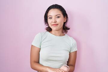 Hispanic young woman standing over pink background with hands together and crossed fingers smiling relaxed and cheerful. success and optimistic