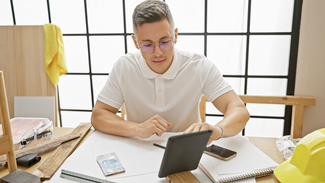 Young hispanic man reviewing plans on a tablet in a carpentry workshop.