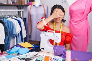 Hispanic young woman dressmaker designer using sewing machine covering eyes with hand, looking...