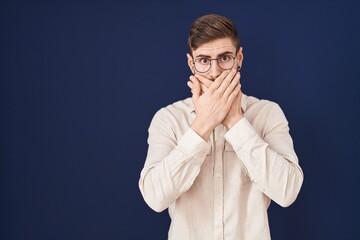 Hispanic man with beard standing over blue background shocked covering mouth with hands for...