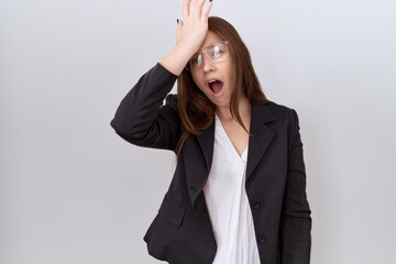 Beautiful brunette woman wearing business jacket and glasses surprised with hand on head for...