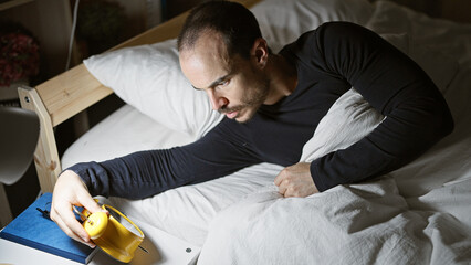 A bald man with a beard, setting an alarm on a yellow clock in a dimly lit bedroom at night.