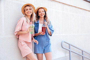 Two young beautiful smiling hipster female in trendy summer clothes. Carefree women posing outdoors. Positive models holding and drinking fresh cocktail smoothie drink in plastic cup with straw