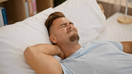 Obraz premium Young hispanic man with a beard showing discomfort in a bedroom setting, lying on white bedding, expressing pain.
