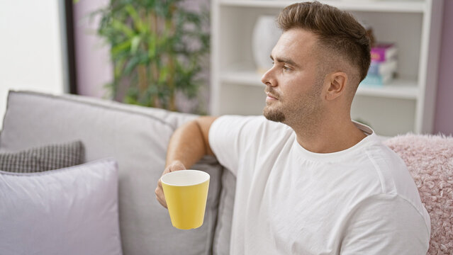Handsome young hispanic man with beard in a white shirt holding a coffee cup, gazing away thoughtfully in a modern living room.