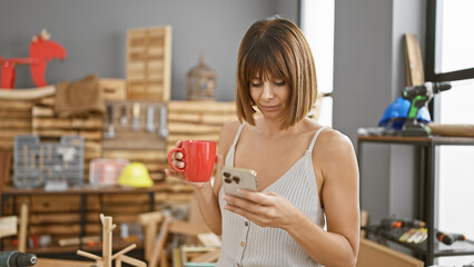 Portrait of a serious young beautiful hispanic woman carpenter, handling her smartphone and savoring a warm cup of coffee in her cozy carpentry workshop.