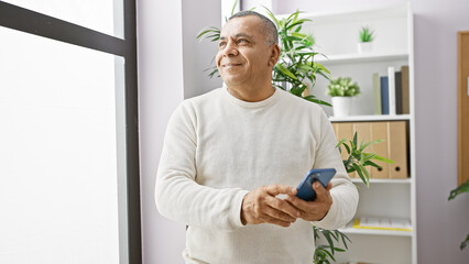 Middle-aged hispanic man in sweater using smartphone in modern office