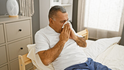 Mature hispanic man sneezing in a modern bedroom setting, capturing a moment of illness or allergy...