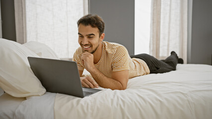 A smiling young man lies on a bed with a laptop in a bright bedroom, indicating comfort and leisure...