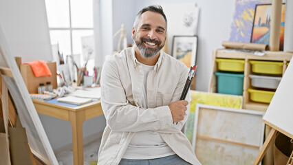 Smiling man with beard holding paintbrushes in art studio indoor setting.