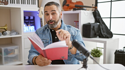 Handsome senior hispanic man with grey beard recording in studio, reading book wearing denim jacket