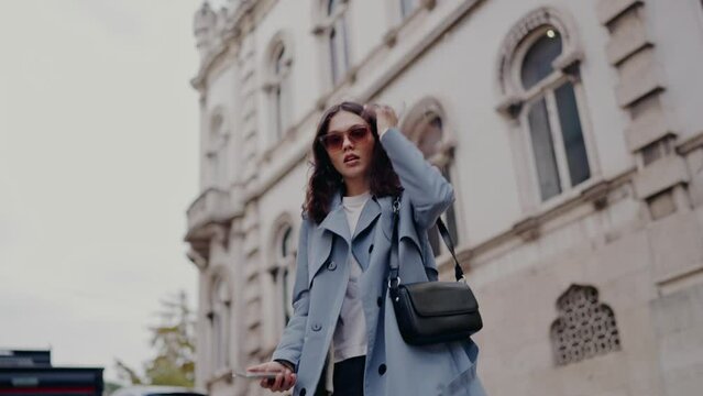 Young woman in stylish outerwear and sunglasses walking on street in city. Pretty female strolling in trench coat with handbag outdoors on architecture background