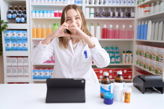 Young caucasian woman working at pharmacy drugstore using tablet smiling in love showing heart symbol and shape with hands. romantic concept.