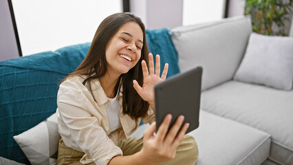 Gorgeous, fun-loving young hispanic woman basks in the joy of a smile-filled video call while comfortably lounging on a sofa at her charming home, radiating confidence and happiness.