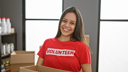 Confident, young hispanic woman smiling brightly, volunteering at community charity center, holding...