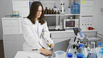 Confident smiling young hispanic woman, a beautiful scientist, working at her laptop in a bustling laboratory, immersed in groundbreaking medical research.