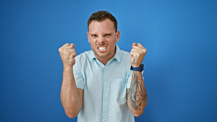 Angry young hispanic man with beard clenched fists against blue wall.