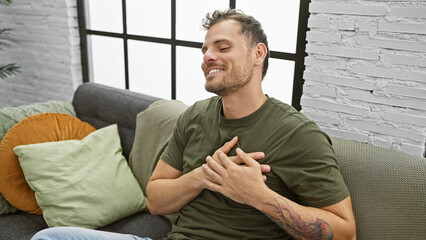 Fototapeta premium A bearded young man sits relaxed at home on a gray sofa, displaying a joyful expression and casual style.