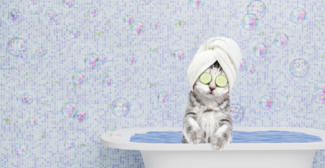 Cute kitten with towel on it head, with pieces of cucumber on it eyes and with cream on it face takes the bath at home. Empty space for text