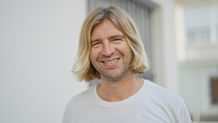 A smiling man with long blond hair posing outdoors on a sunny urban street.