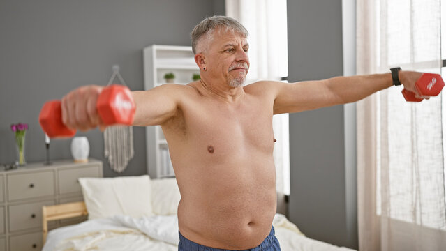Middle-aged shirtless man exercising with dumbbells in a modern bedroom, maintaining fitness at home.