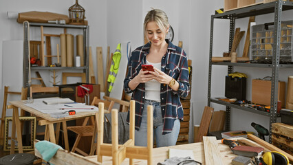 A young woman smiles while using her phone in a cluttered woodworking workshop.