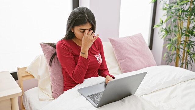 A stressed indian woman with long hair sits on a bed with a laptop in a home bedroom, eliciting emotions of exhaustion or concern.