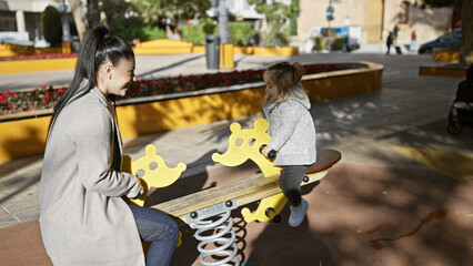 A young woman and little girl enjoy playful bonding on a seesaw at a sunny outdoor playground,...