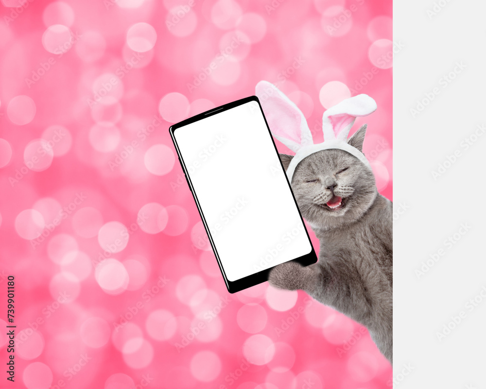 Poster happy kitten wearing easter rabbits ears holds big smartphone with white blank screen and looks from
