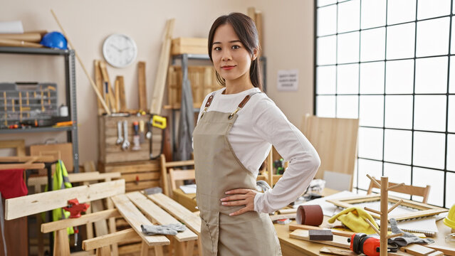 A confident young asian woman posing in a well-equipped carpentry workshop, brimming with tools and timber.