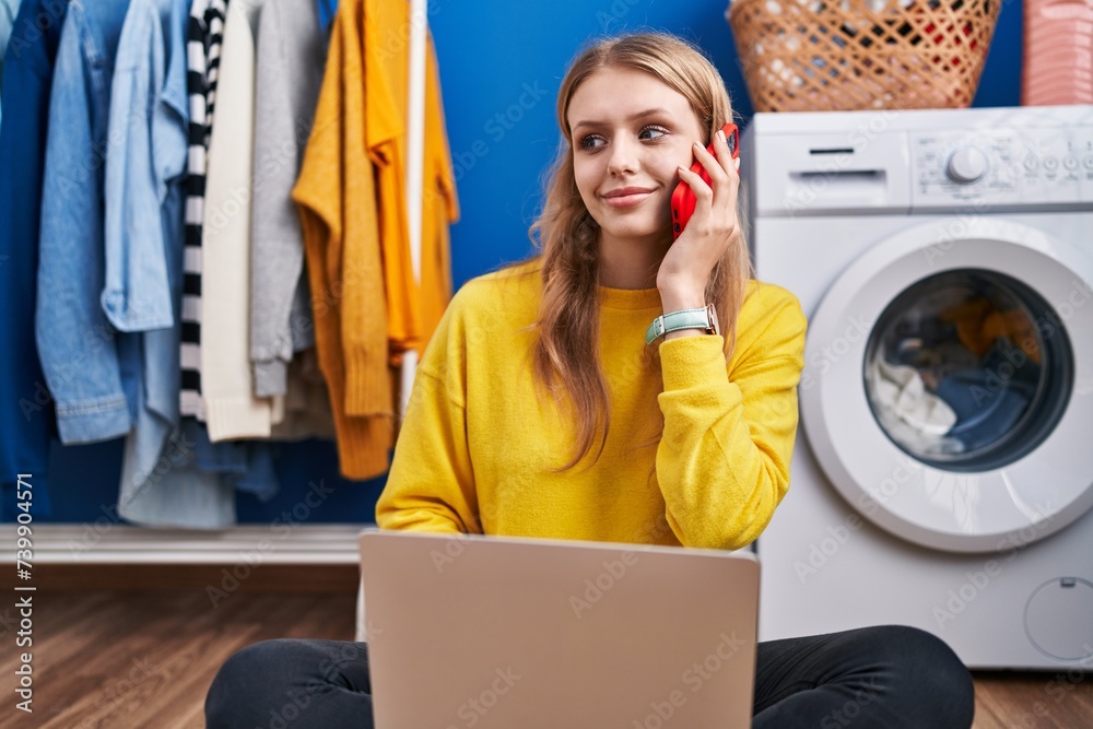 Wall mural young blonde woman using laptop talking on smartphone waiting for washing machine at laundry room