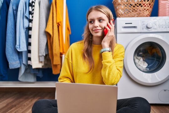 Young blonde woman using laptop talking on smartphone waiting for washing machine at laundry room
