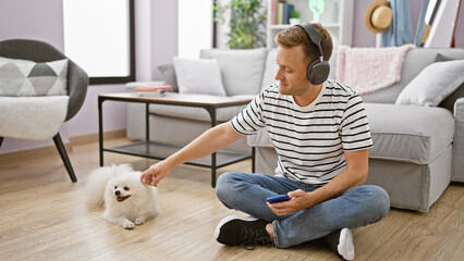 Engrossed young caucasian man sitting on the floor at home, deeply focused on a song playing on his smartphone using headphones while his adult dog patiently waits beside him - Powered by Adobe