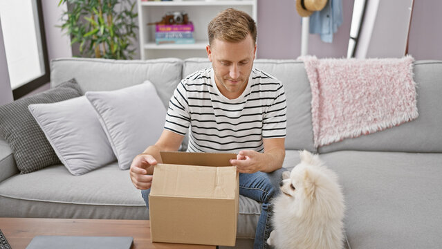 Intrigued young caucasian man patiently unpacking cardboard box delivery at home, his curious dog perched nearby, both ensconced in their cozy apartment living room background.